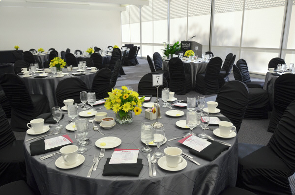 Several grey linen covered tables and black linen covered chairs with crystalware, porcelain plates, and silverware and yellow flower centerpieces.